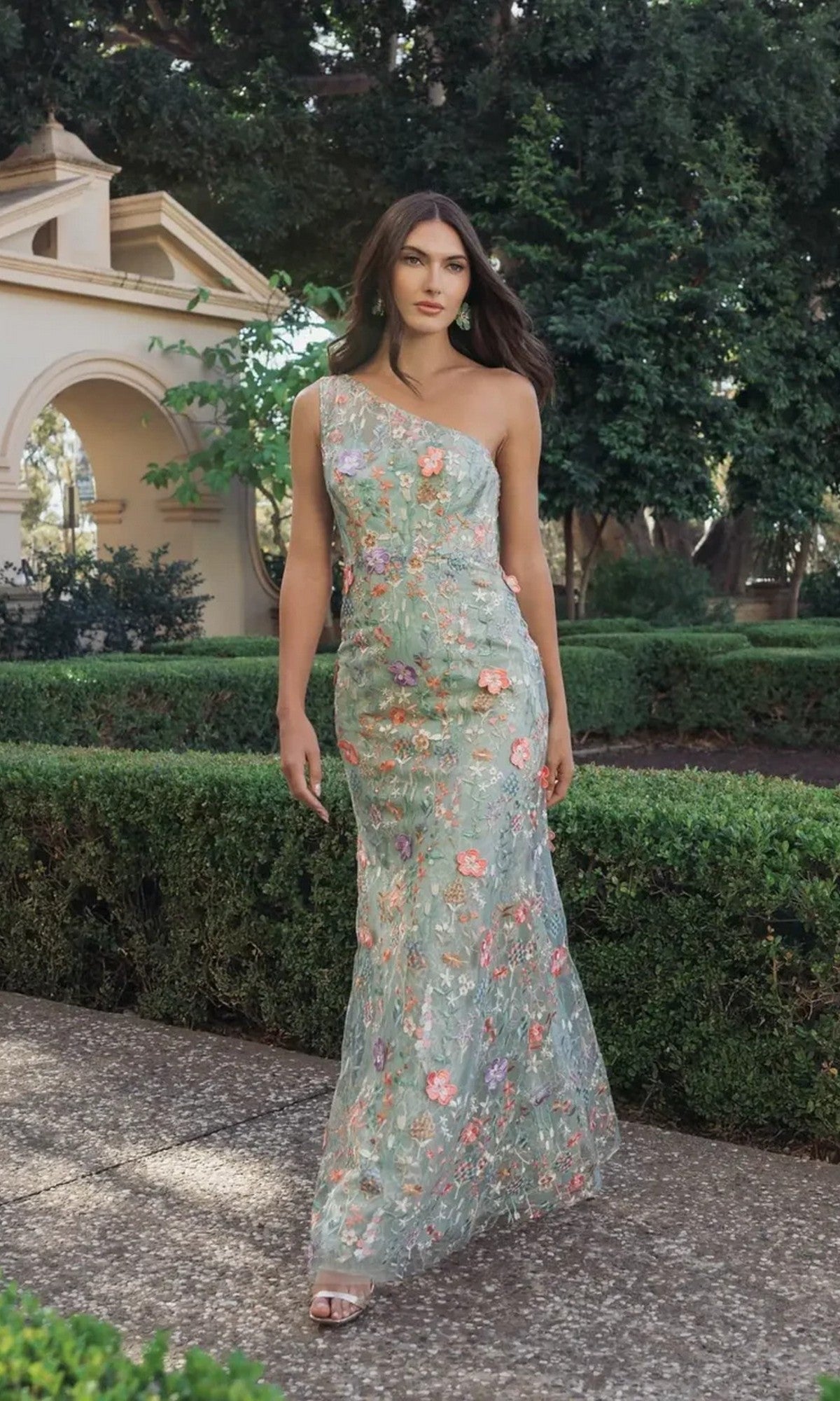 A woman in the Mac Duggal 68606 Long Floral-Print Formal Dress in Sage Multi strolls along a garden path, surrounded by lush hedges and trees.